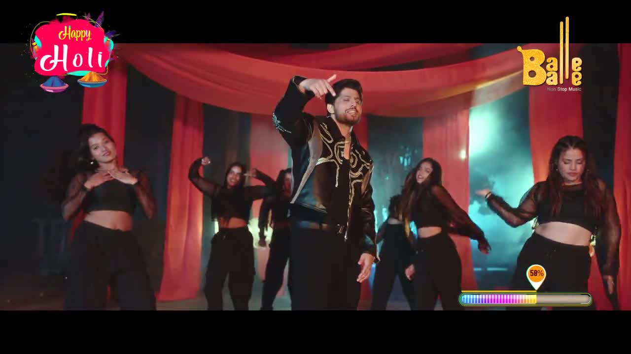 A man in a black jacket with gold trim moves his hands in a sharp gesture, surrounded by women in matching black outfits. The backdrop is a stage with red drapes, hinting at a festive Indian celebration.