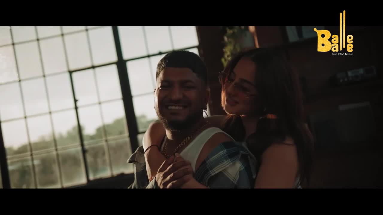 A man, smiling broadly, has his arms around a woman who leans her head on his shoulder. Sunlight streams through large windows behind them.