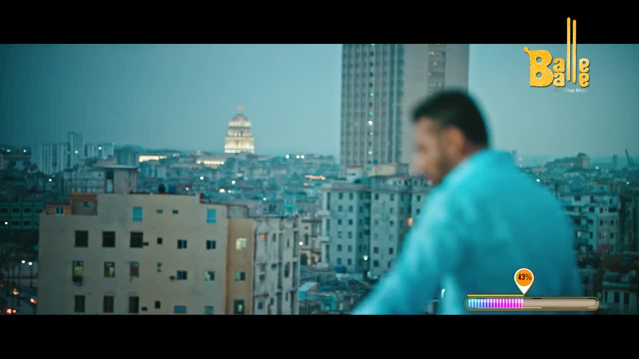 A man in a bright blue shirt stands on a rooftop, looking out over a city at dusk. Buildings stretch into the hazy sky, punctuated by a distant, illuminated dome.