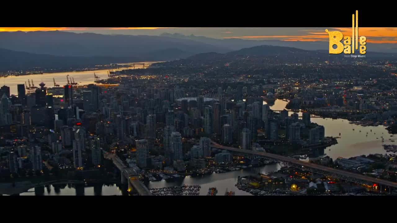 The city lights twinkle as the sun dips below the hazy mountains. Boats dot the water, reflecting the fading glow of dusk.