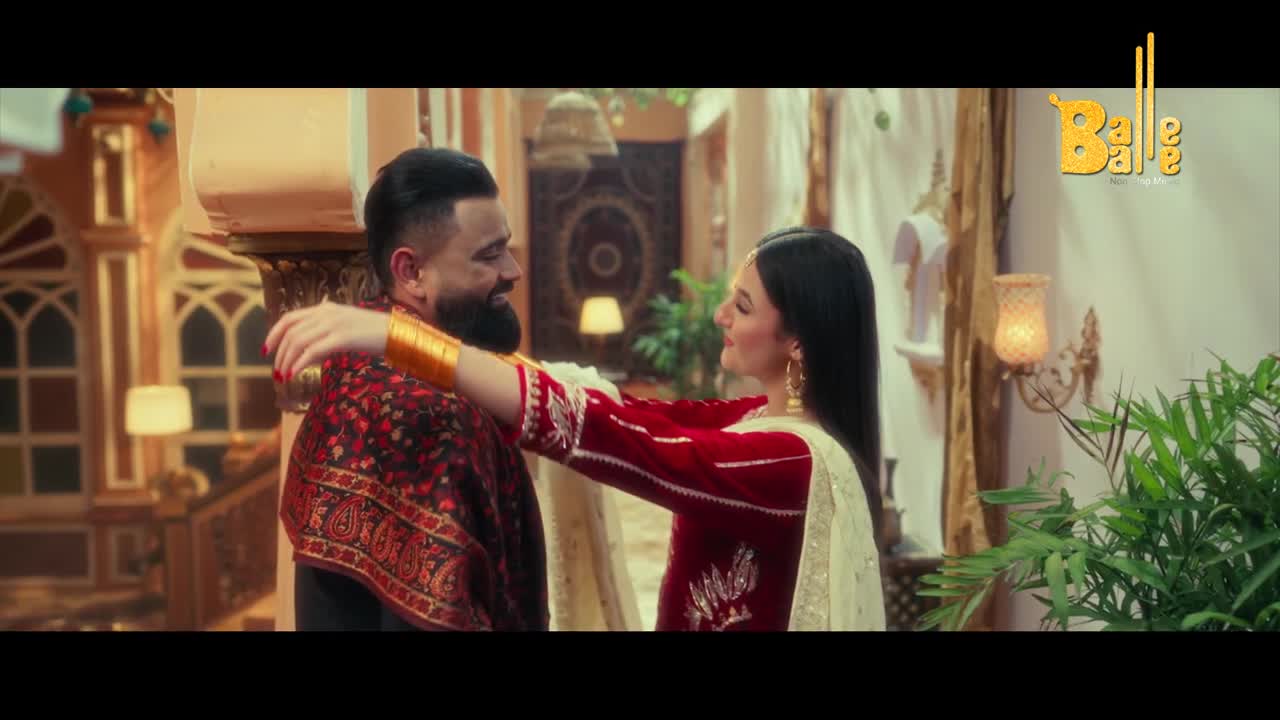 A woman in a red outfit, adorned with gold bangles, embraces a man wearing a patterned shawl. They share a warm smile in a richly decorated room.
