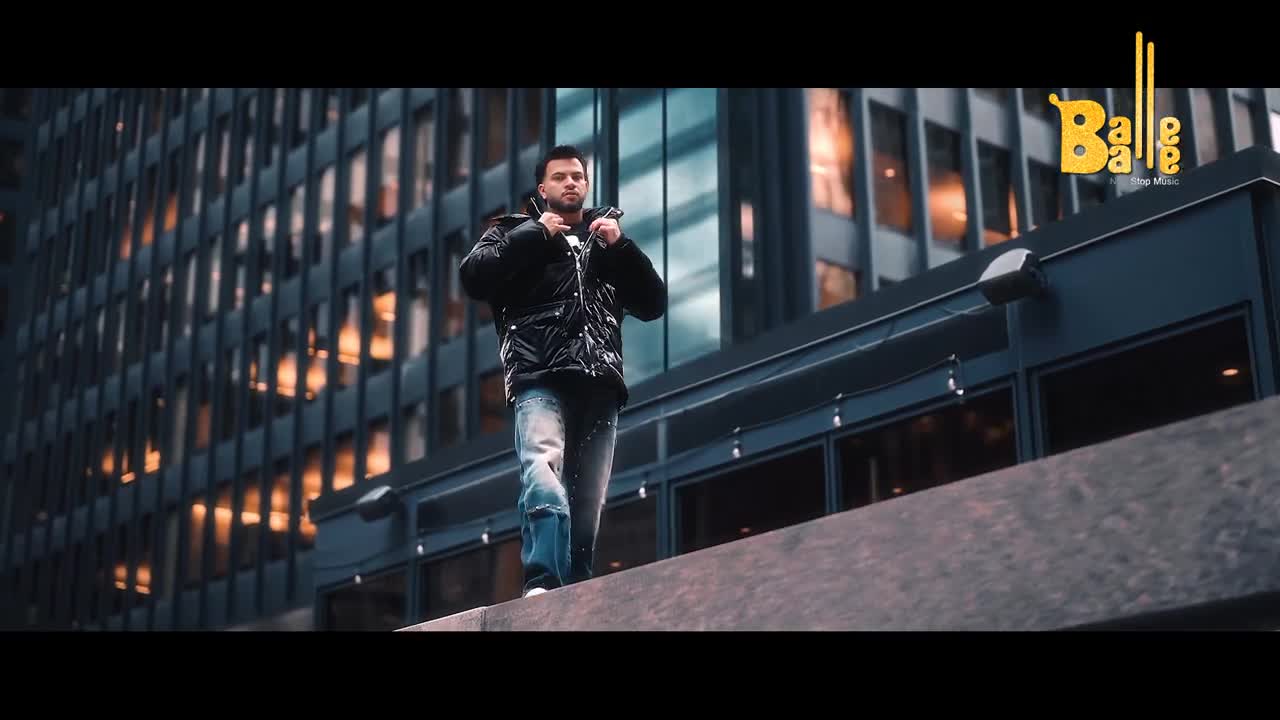 A man walks along a ledge, his black jacket shimmering against the backdrop of a modern building. The "Balle Balle" logo appears in the upper right corner, hinting at a Punjabi music video.