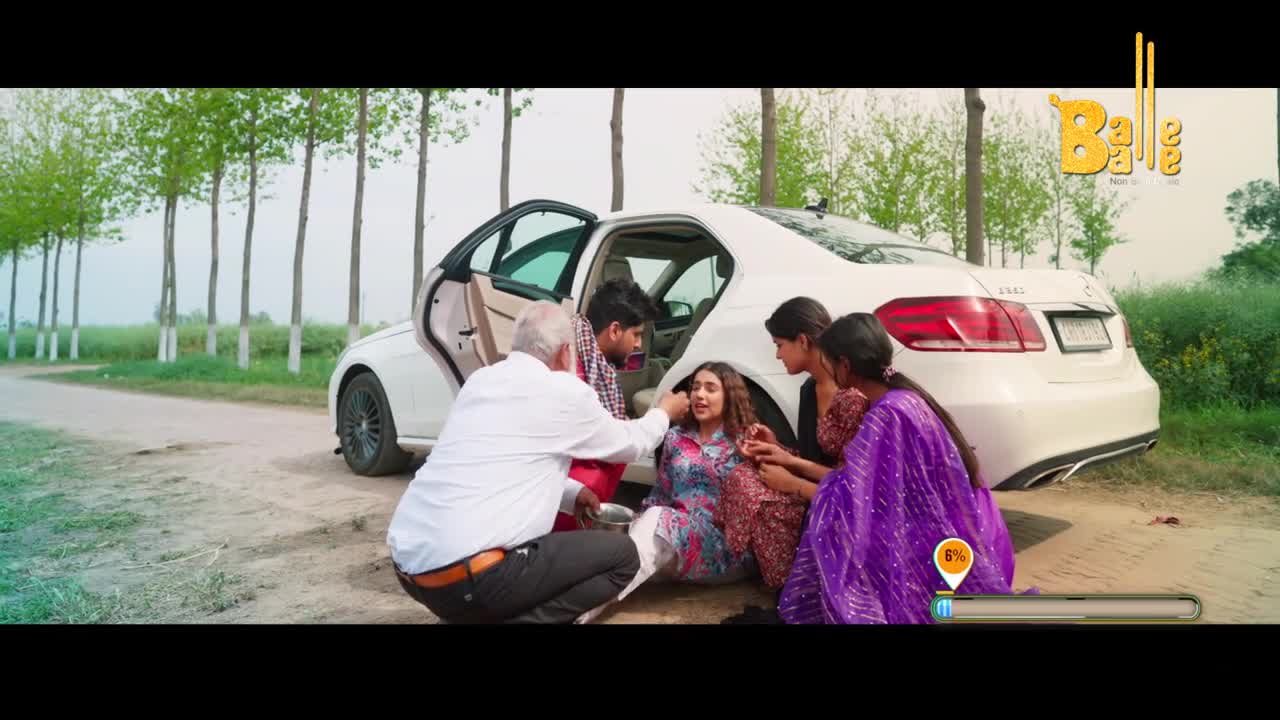 A young woman is being helped up from the ground next to a white car. Two women in colorful saris and an older man in a white shirt surround her, offering support.