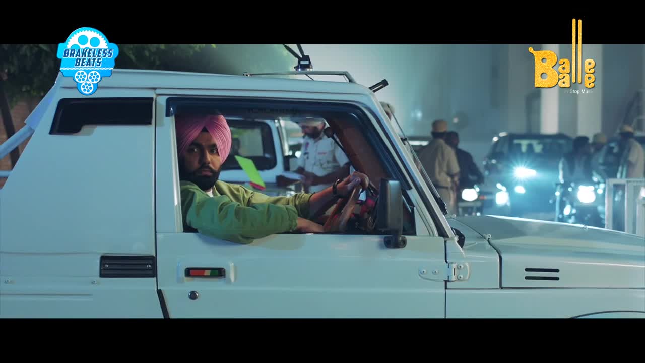 A man in a pink turban grips the steering wheel of a white jeep. Behind him, several figures in uniform stand near other vehicles under bright lights.