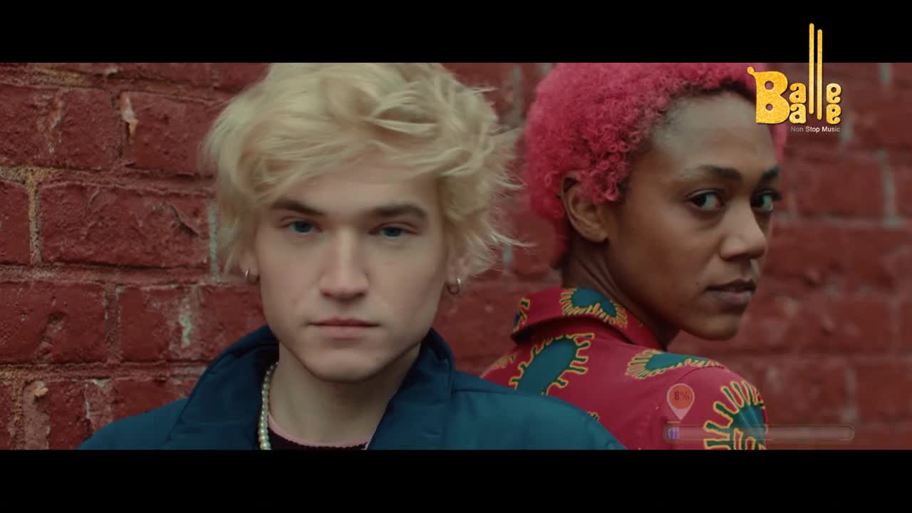 A young man with bright blonde hair stands close to a red brick wall, his gaze direct. Behind him, a woman with vibrant pink hair turns her head, her expression thoughtful.