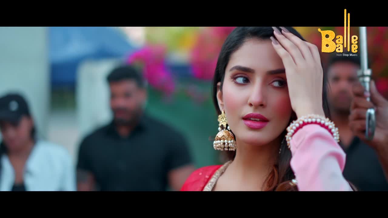 A woman adjusts her large, pearl-studded jhumka earrings, her eyes glancing sideways. Behind her, two men in dark shirts stand, one with a blurred face, as colorful decorations hang in the background.