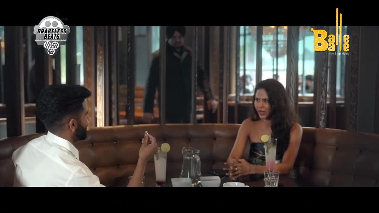 A man in a dark green kurta and turban walks past a booth where a woman in a strapless top is talking animatedly. Another person, seen from behind in a white shirt, sits opposite her, holding a small object.