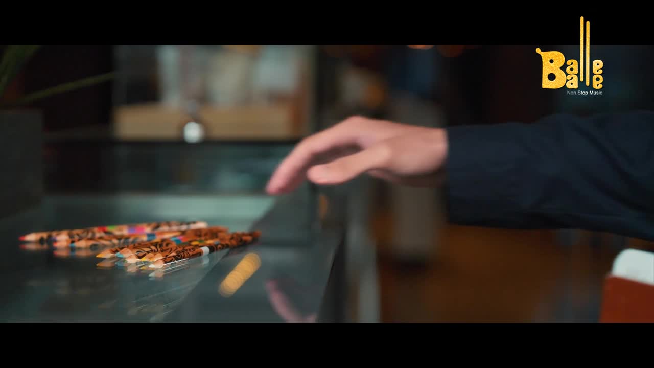 A hand hovers over a line of colorful pencils and coffee beans arranged on a glass surface. The Balle Balle logo appears in the upper right corner.