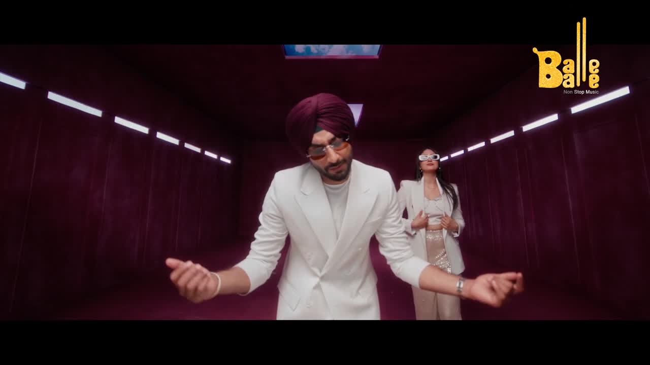 A man in a white suit and maroon turban moves his hands with a flourish. Behind him, a woman in a similar suit stands, looking towards the camera.