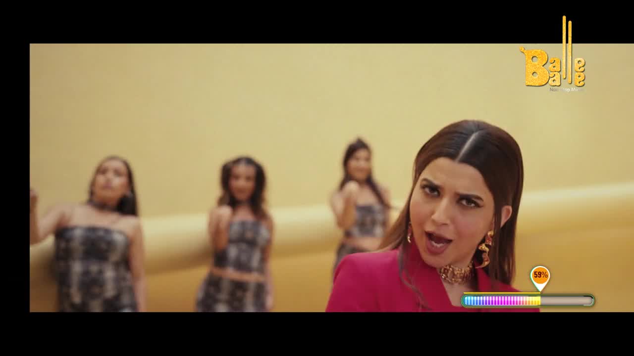 A woman in a bright pink jacket sings into the camera, her mouth open. Behind her, three other women in matching patterned outfits sway to the music.