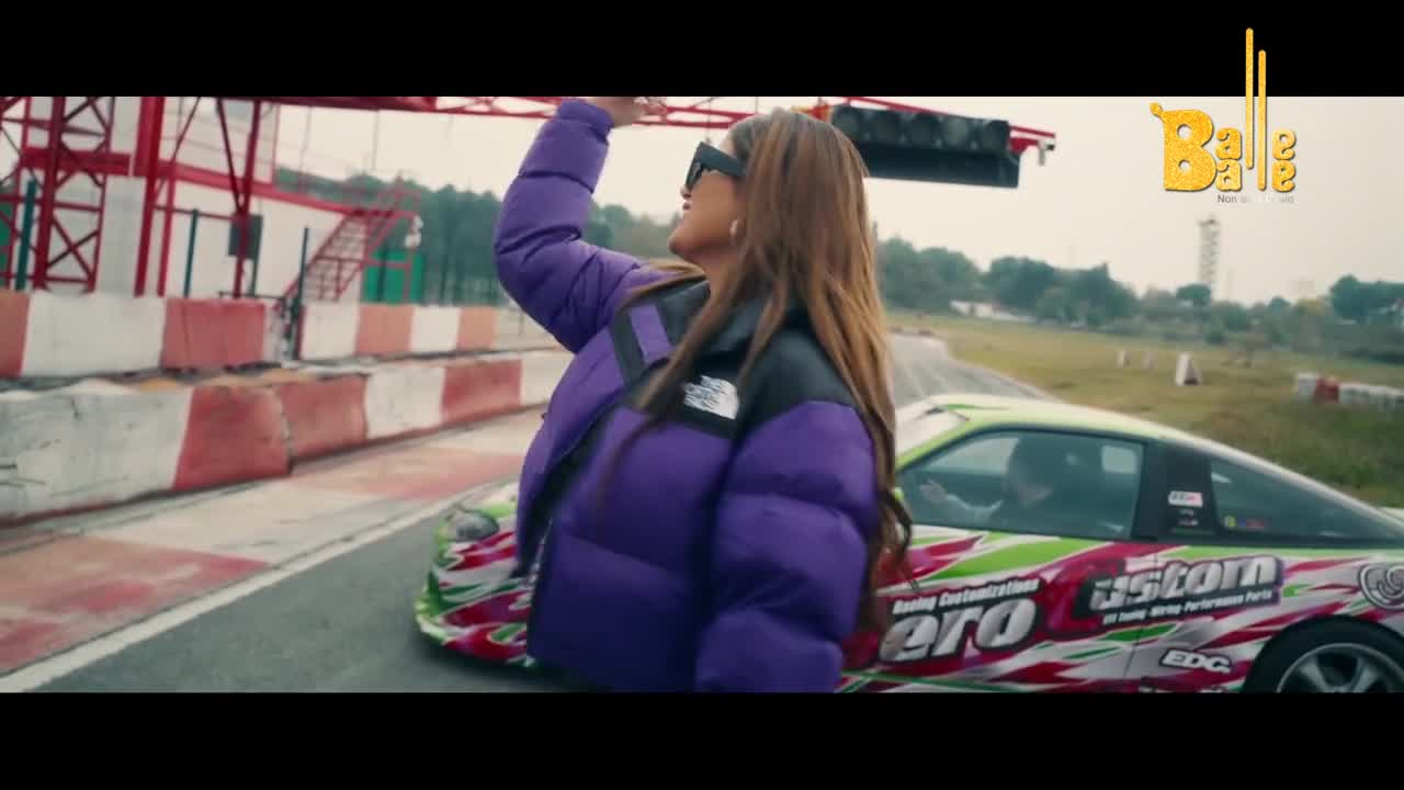 A bright green car with a wild graphic design slides around the track. A woman in a puffy purple jacket stands nearby, her arm raised.