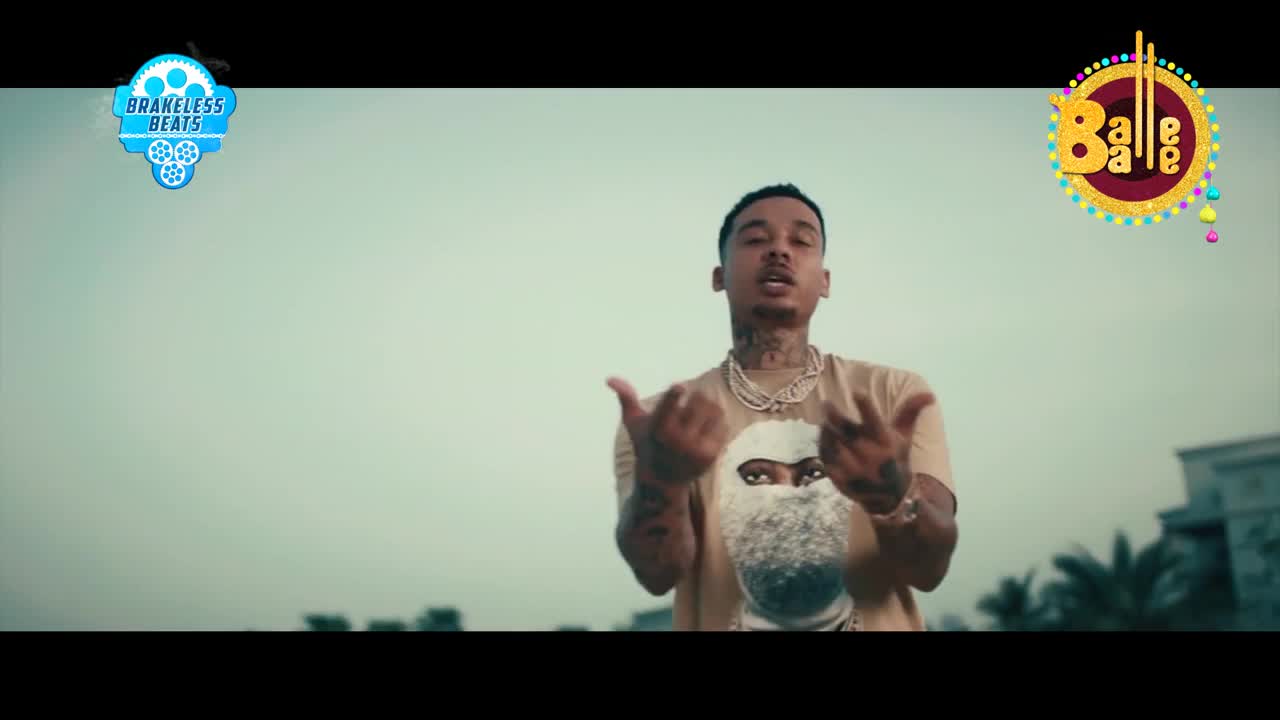 A young man with tattoos gestures with his hands, his face partially obscured by a graphic on his shirt. Buildings and palm trees are visible in the background under a pale sky.