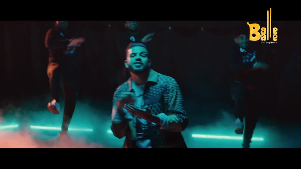 A man with a patterned shirt sings into the camera as dancers in dark clothing move around him. Teal light illuminates the smoky stage, a common scene for a Balle Balle production.
