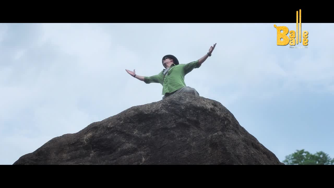 A man in a green shirt and hat stands on a large rock, arms outstretched towards the sky. The Balle Balle logo appears in the upper right corner.