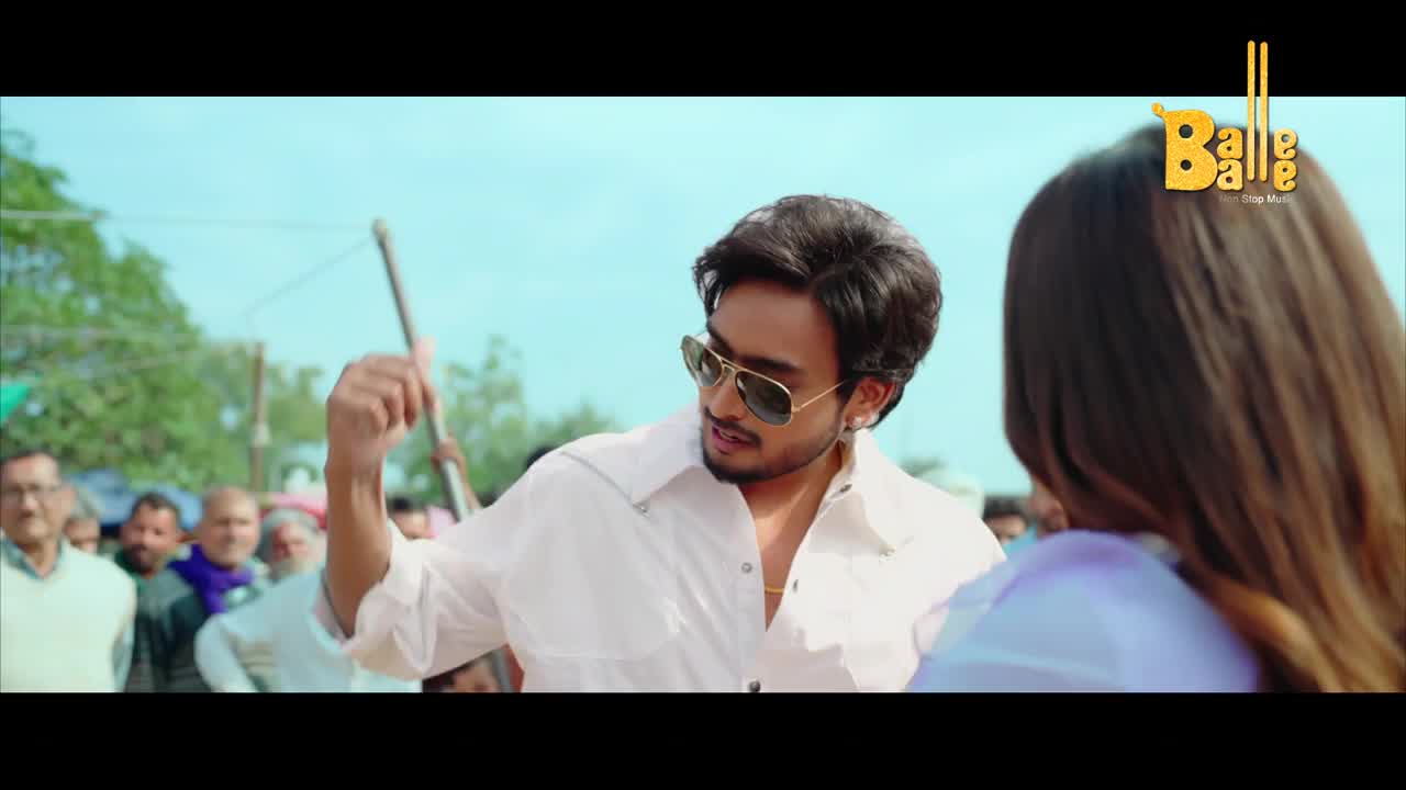 A young man in a white shirt and sunglasses gestures with his right hand, a stick held behind him. A crowd of people, some in traditional Indian attire, watches the scene unfold under a bright sky.