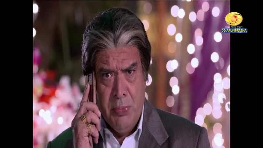 The man in the suit holds a phone to his ear, his brow furrowed in concentration. The DD Arun Prabha logo in the corner suggests this is a scene from a show.
