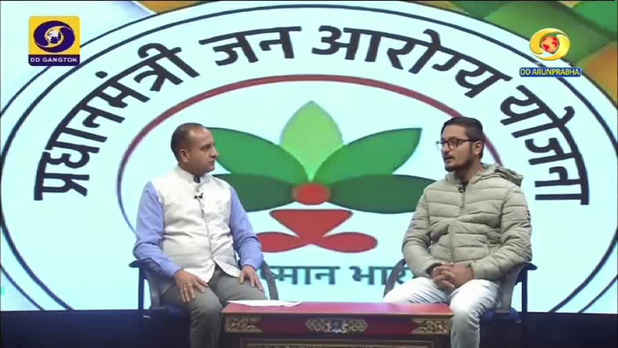 Two men sit on a stage in front of a large logo, the DD Arun Prabha logo visible in the corner. The man on the right, in a light jacket, is speaking while the other listens intently.
