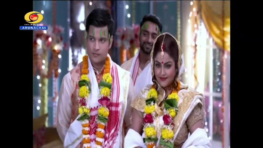 A couple, adorned with garlands, stands together, a man smiling behind them. The scene, likely from a DD Arun Prabha broadcast in India, seems to be a wedding ceremony.
