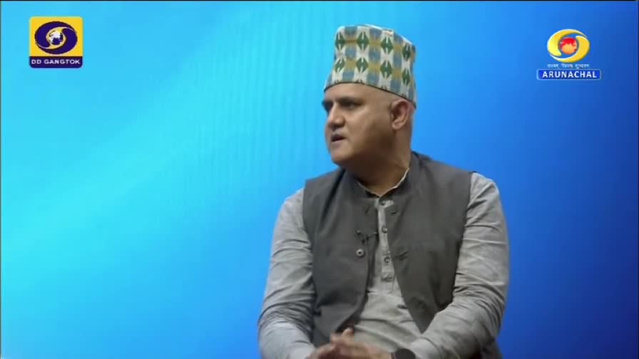 The man in the patterned hat is speaking, his gaze directed off-camera. The DD Arun Prabha logo is visible in the background, suggesting a broadcast from India.
