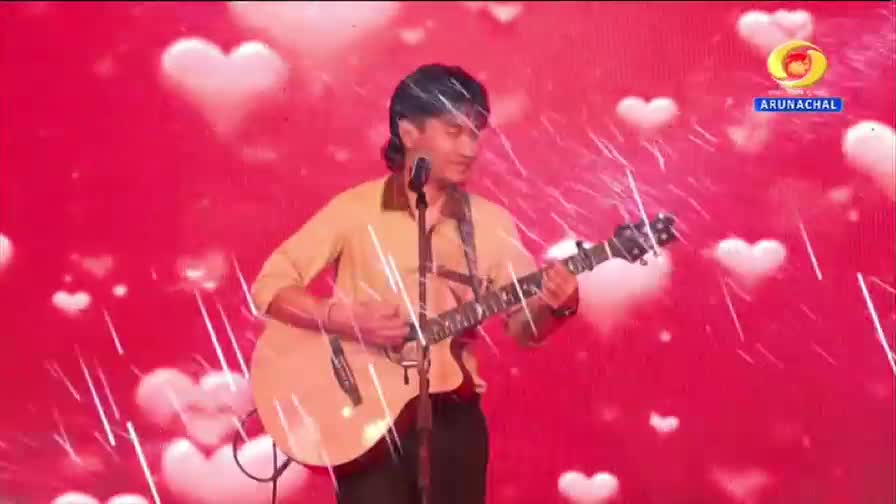 A man strums a guitar on a stage, lit by a red backdrop dotted with white hearts. Sparkling effects burst around him as he sings, likely on DD Arun Prabha from India.
