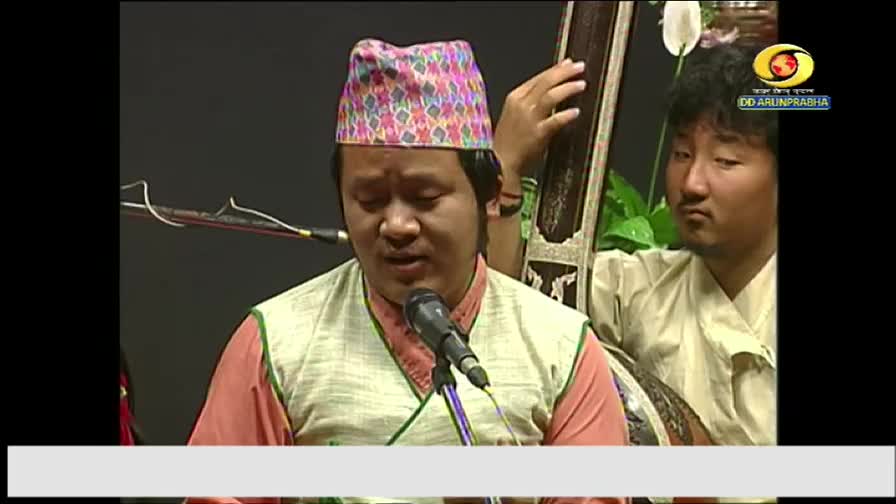 The singer, wearing a patterned hat, is singing into a microphone, his face animated. Beside him, a musician plucks at a stringed instrument, the DD Arun Prabha logo visible in the corner.
