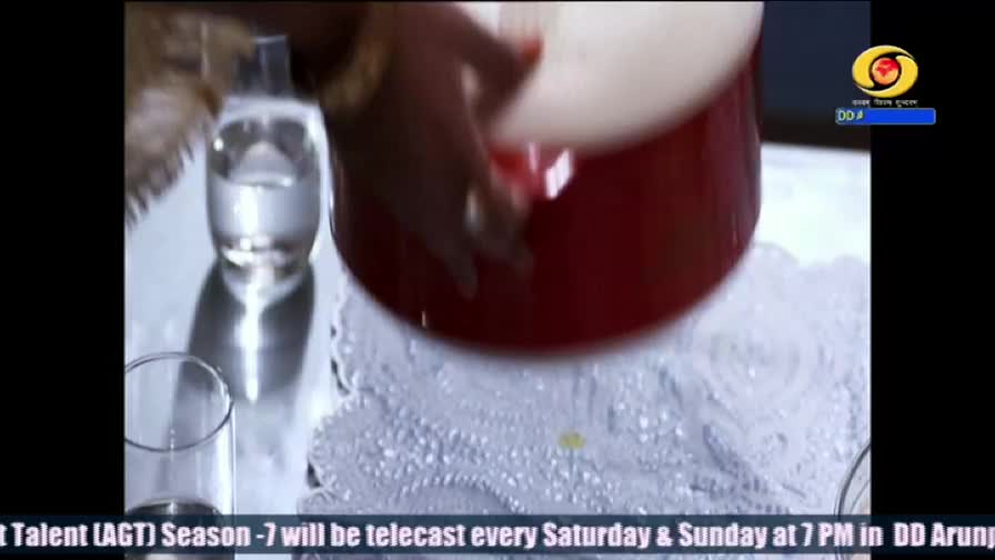 A hand, adorned with a gold bracelet, is holding a large red pot. The pot is being lowered onto a white, textured surface, as seen on DD Arun Prabha.
