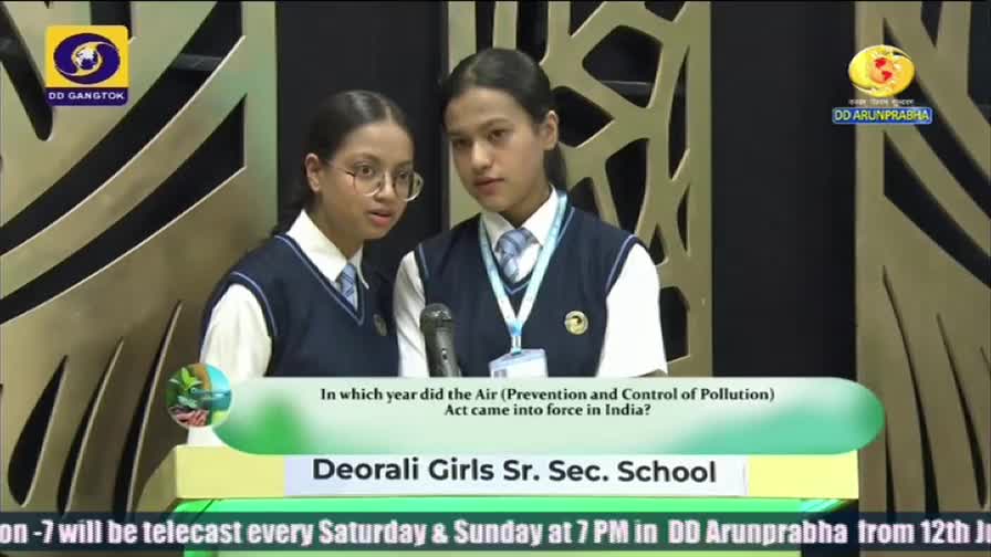 Two students from Deorali Girls Sr. Sec. School stand at a podium, facing the camera, with a microphone between them. The DD Arunprabha logo is visible, and a question about an Indian environmental act is displayed on the screen.
