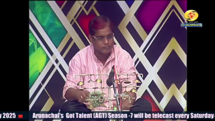 A man in glasses sits cross-legged, holding a small instrument with a series of bells. He seems focused, ready to play on this DD Arun Prabha program from India.
