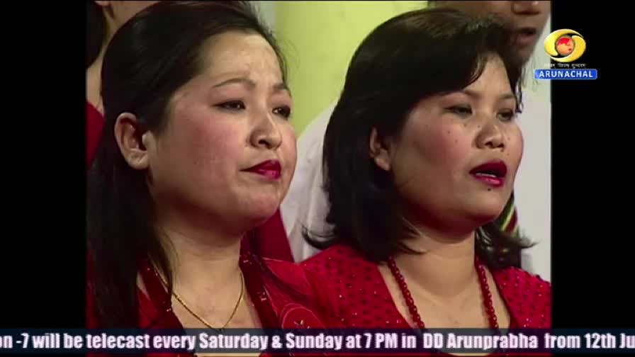 Two women in red tops are singing, their mouths open as they perform. The DD Arunprabha logo is visible in the corner, and the broadcast details are scrolling at the bottom.
