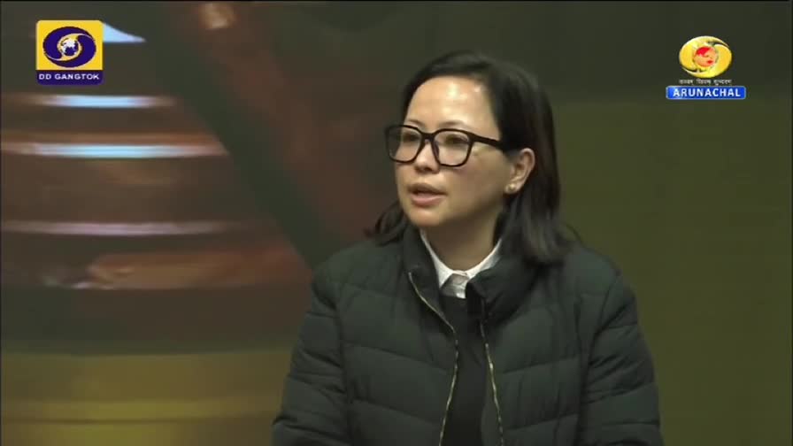 A woman in glasses, wearing a black jacket, speaks on a DD Arun Prabha broadcast. The DD Gangtok logo sits in the corner.
