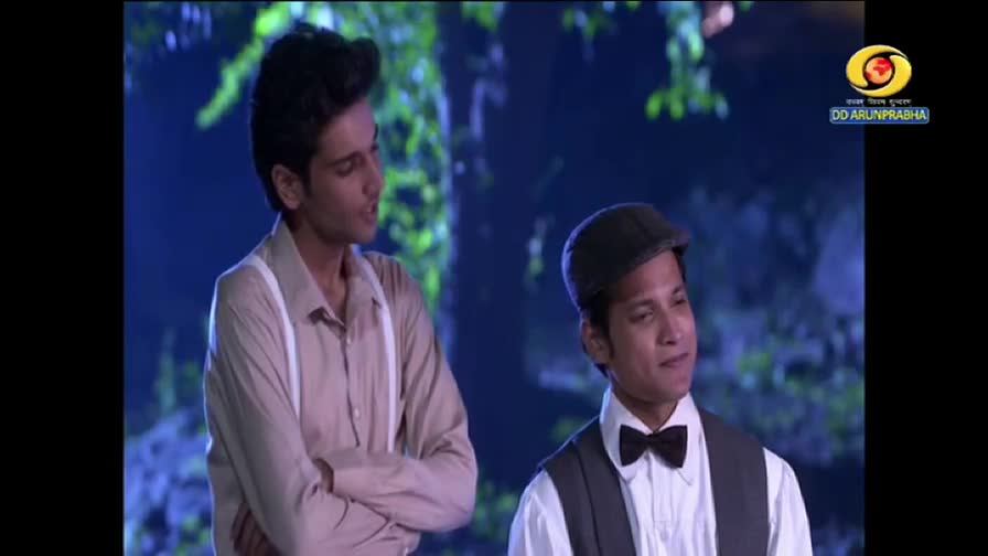 Two men, one in a cap and bow tie, are on screen, likely from a DD Arun Prabha show. The other man, in a light shirt and suspenders, seems to be speaking to the other.
