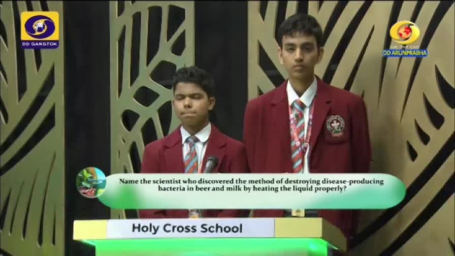 Two students in maroon blazers stand behind a podium, ready to answer a question posed on the DD Arun Prabha quiz show. The question, displayed on a green banner, asks about a scientist's discovery.
