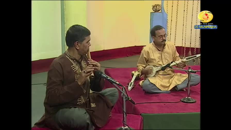 Two men sit cross-legged on a red platform, one playing a flute, the other a stringed instrument. The DD Arun Prabha logo sits in the corner, and the scene is likely a performance from India.
