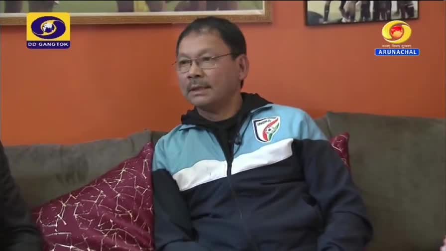 A man in glasses and a blue and black jacket is seated on a couch, likely being interviewed. The DD Arun Prabha logo is visible in the background, suggesting this is a broadcast from India.
