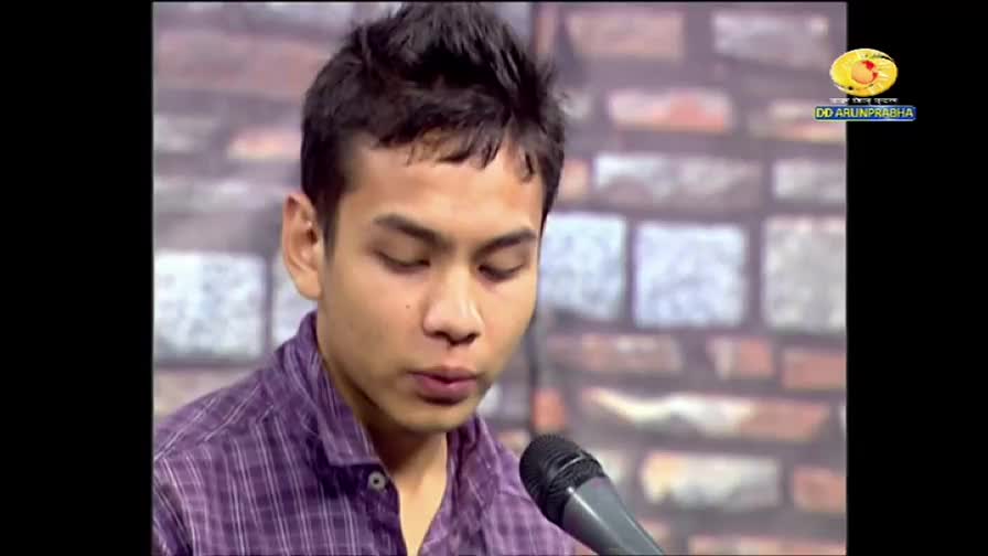 A young man in a purple plaid shirt speaks into a microphone, his eyes downcast. The DD Arun Prabha logo is visible in the corner, suggesting a broadcast from India.
