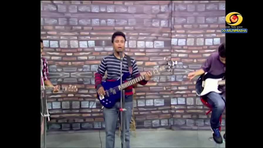 A band is playing on a stage with a brick wall backdrop, likely for a DD Arun Prabha broadcast. The singer, wearing a striped shirt, strums a blue bass guitar, while the others play alongside him.
