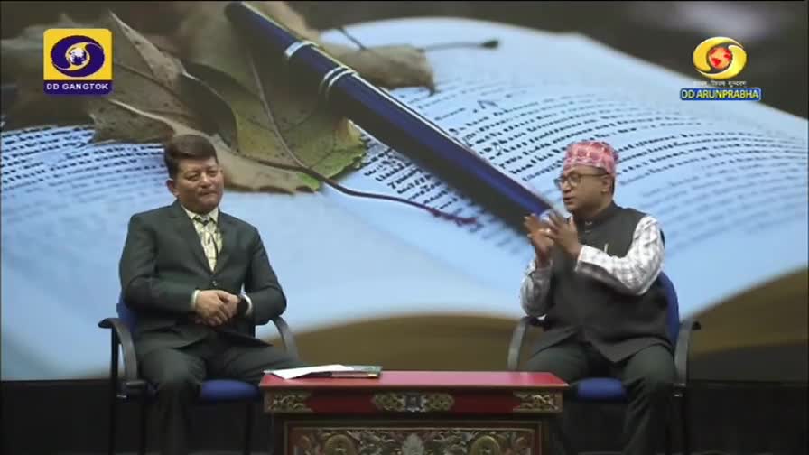 Two men are seated, facing each other in a studio for DD Arun Prabha, with a backdrop of a book and pen. The man on the right gestures with his hands, while the other listens intently.
