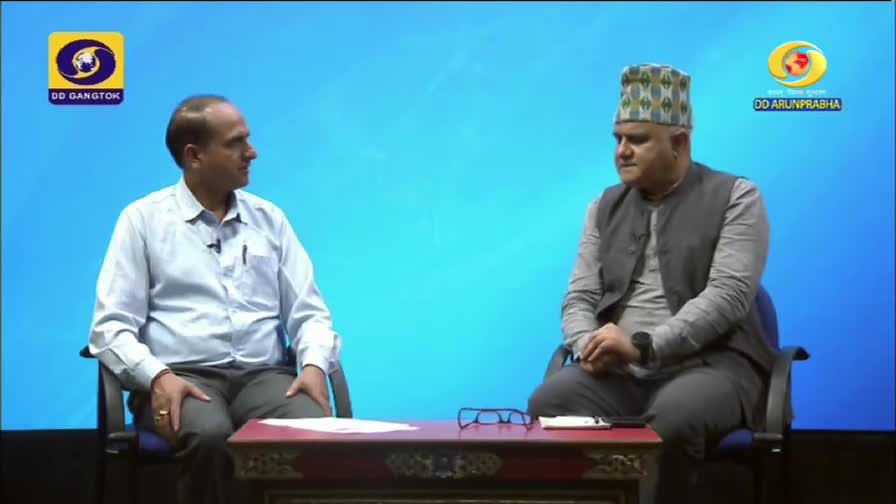 Two men sit facing each other on a set for DD Arun Prabha, with a small table between them. One wears a patterned cap and a vest, while the other wears a button-down shirt.
