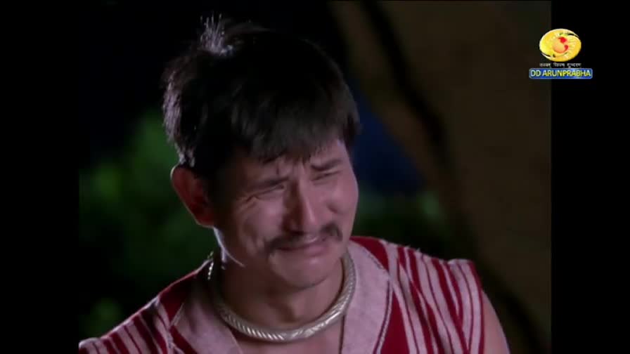 The man is squinting, his face contorted with emotion, perhaps sadness. His red and white striped shirt and the DD Arun Prabha logo suggest a broadcast from India.
