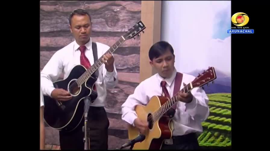 Two men, both in white shirts and ties, strum acoustic guitars on a DD Arun Prabha set. The backdrop is a simple painted landscape, with the Arunachal Pradesh logo visible.

