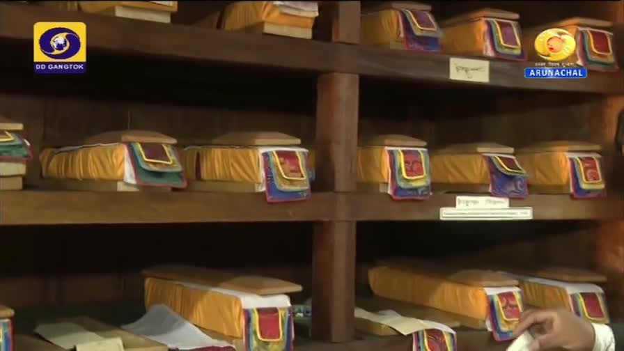 Wooden shelves hold rows of rectangular boxes, each covered in yellow fabric and adorned with colorful trim. The DD Arun Prabha logo in the corner suggests a broadcast from India.
