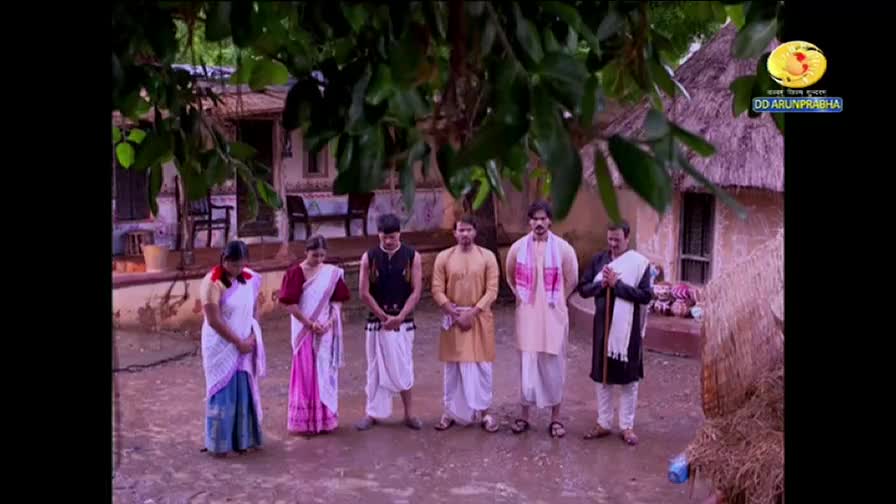 A group of people stand in a line, heads bowed, in what appears to be a rural setting. The scene, broadcast on DD Arun Prabha, suggests a moment of respect or reflection.
