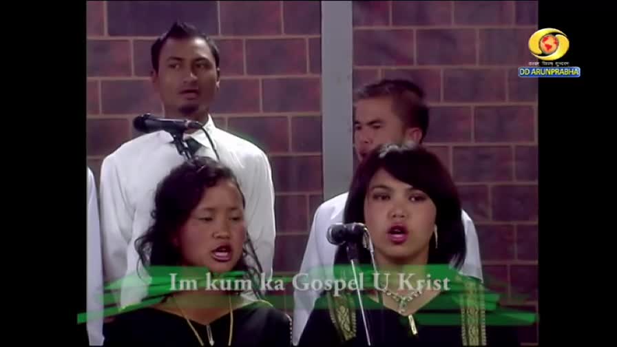 A group of people, dressed in white shirts, sing into microphones on a stage. The DD Arun Prabha logo is visible, and the words "Im kum ka Gospel U Krist" are displayed on the screen.

