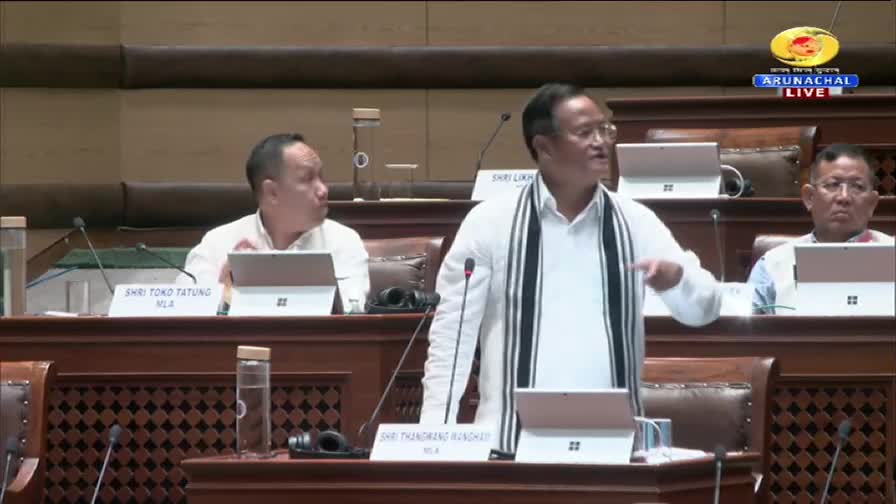 A man in a white shirt and striped shawl gestures emphatically towards the right, speaking into a microphone. Two other men sit at desks, watching the proceedings, as the Arunachal Live broadcast continues on DD Arun Prabha.
