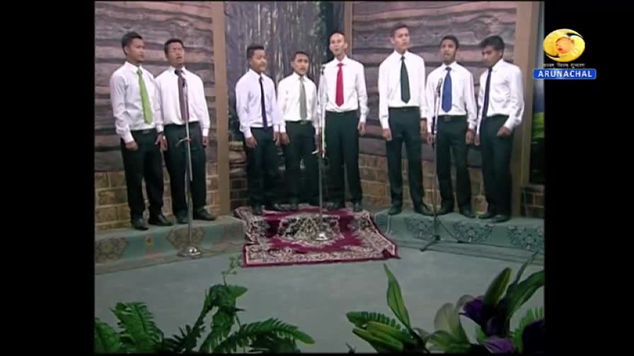 Seven men in white shirts and ties stand in a row, each facing a microphone. They're on a small stage, with a patterned rug at their feet, likely performing on DD Arun Prabha.
