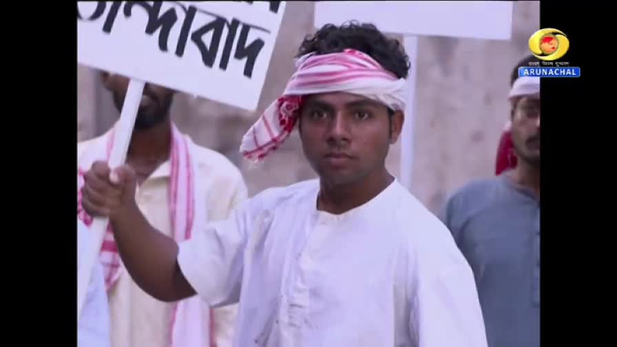 A young man, wearing a white kurta and a patterned headscarf, holds a sign aloft, his gaze fixed forward. Behind him, other men in similar attire stand in a line, suggesting a protest scene, possibly from a DD Arun Prabha broadcast in India.
