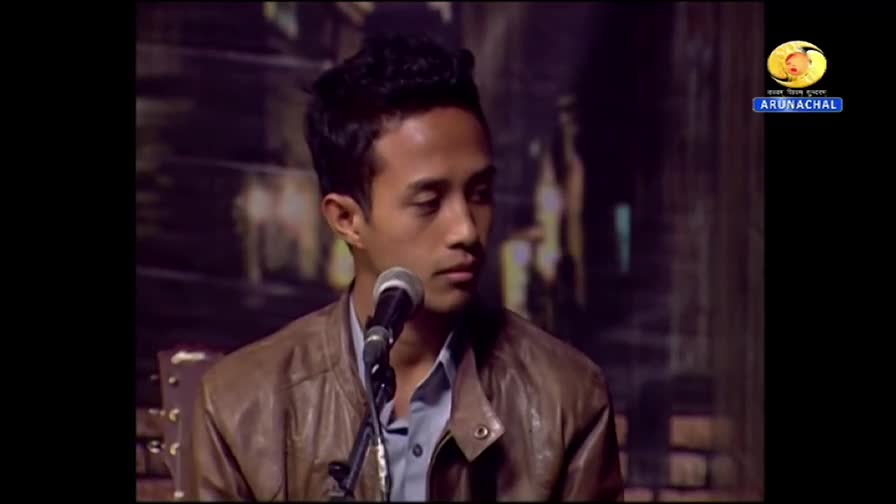 A young man in a brown leather jacket is speaking into a microphone, his gaze fixed downwards. The DD Arun Prabha logo is visible in the corner, indicating this is likely a broadcast from India.
