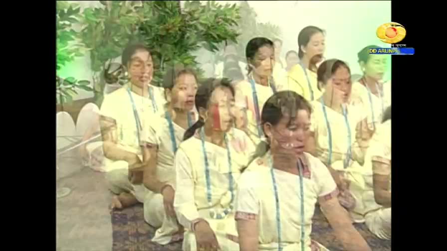 A group of women in white tunics and blue necklaces sit close together, some with red markings on their faces. They appear to be singing, their mouths open in unison, as the DD Arun Prabha logo is visible in the corner.
