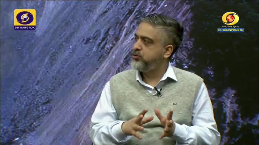 A man in a light sweater gestures with his hands as he speaks on a DD Arun Prabha program. The backdrop is a textured, dark blue surface, and the DD Gangtok logo is visible in the corner.
