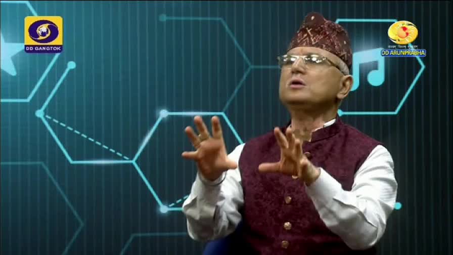 A man in a patterned hat and vest gestures with both hands, speaking on a set for DD Arun Prabha. The background is a teal screen with a geometric design.
